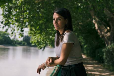 A woman leans on a railing in the foreground, looking out at a calm body of water bordered by trees in the background.