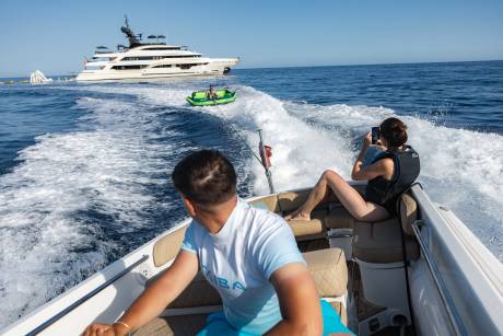 In the foreground, two people are enjoying a ride on a speedboat, while in the background, a large yacht sails on the ocean.