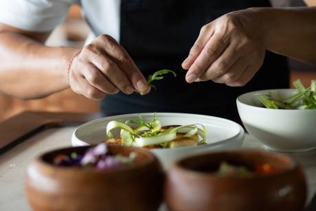 Hands carefully garnish a dish with greens in the foreground, while a blurred kitchen background suggests a professional culinary setting.