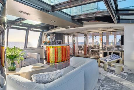 A luxurious yacht interior with plush seating in the foreground and a bar and dining area visible through glass doors in the background, all under a glass ceiling with ocean views.