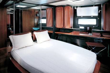 A neatly made bed with white linens dominates the foreground, while the background reveals a compact cabin with mirrored walls, leather storage compartments, and a desk area.