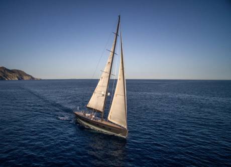 A large sailboat with full sails glides across a deep blue ocean, with a distant, rocky coastline under a clear sky.