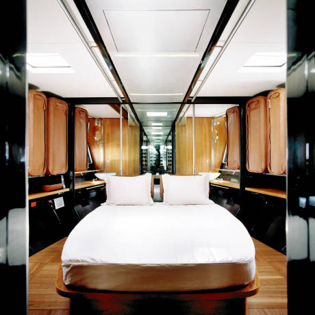 A white-sheeted bed with two pillows sits in the foreground of a luxurious, mirrored room with wooden cabinets lining the walls and a bright ceiling.