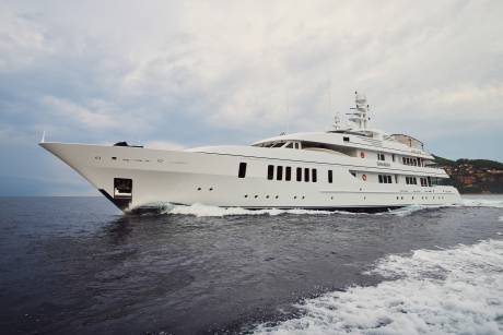 A large white yacht cruises through dark, choppy water with a green, tree-covered hillside in the background under a cloudy sky.
