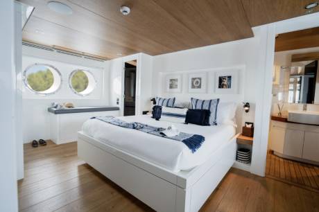 A luxurious bedroom with a large white bed in the foreground and a bathroom with sinks and wooden floors in the background.