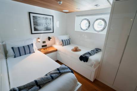 Two neatly made beds with striped pillows and blankets fill the foreground, while two porthole windows and a framed picture adorn the white walls in the background.