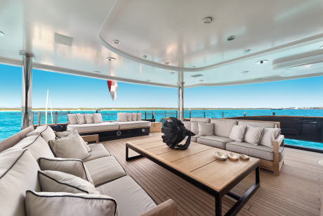 Plush seating and a wooden coffee table fill the foreground of a yacht deck, while the background reveals a bright blue ocean under a clear sky.