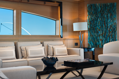A modern living room with plush sofas and a dark coffee table in the foreground, and large windows overlooking a bright blue sky in the background.