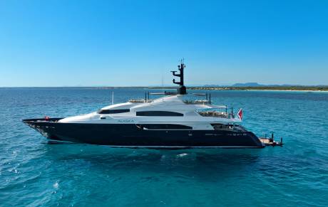A large, dark-hulled yacht named "ALASKA" floats on turquoise water in the foreground, with a distant, hazy coastline under a clear blue sky in the background.