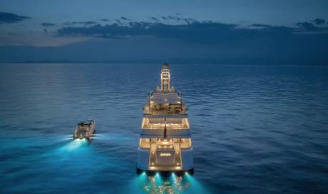 A large yacht and a tender boat are illuminated in the calm, dark ocean under a twilight sky.