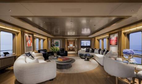 Plush white sofas and armchairs surround a circular rug in the foreground, while large windows offer views of the ocean in the background.