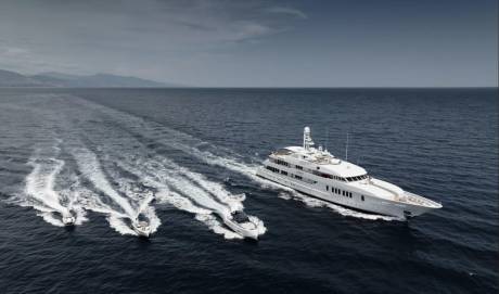 A large white yacht cruises through dark blue water, leaving a wake, with several smaller boats trailing behind it, and a hazy coastline visible in the distance under a cloudy sky.