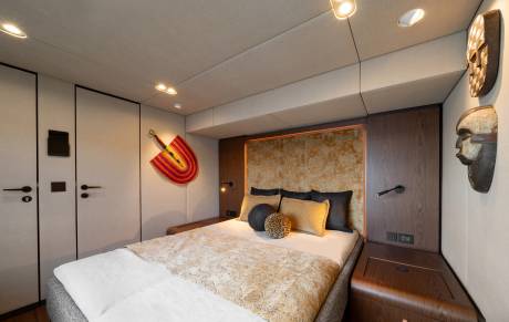 A luxurious bedroom features a plush bed with decorative pillows in the foreground, while tribal masks adorn the wall in the background.
