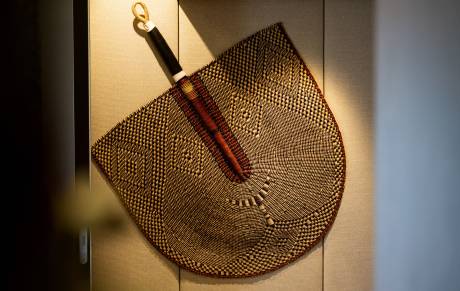 A woven fan with a dark handle and gold loop hangs against a textured, light-colored wall.