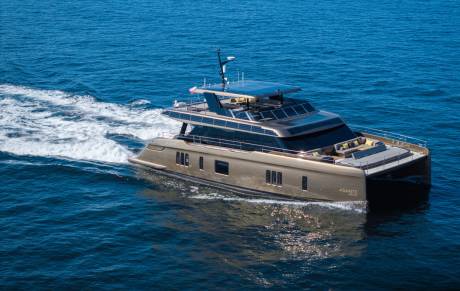 A sleek, golden catamaran yacht glides through calm blue water, leaving a white wake behind it, with a clear sky visible in the distance.