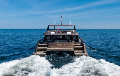A modern catamaran yacht named "ASANTE" cruises through calm blue waters, leaving a white wake behind it under a clear sky.