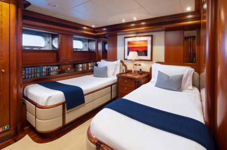 Two neatly made twin beds with blue runners and white linens are in the foreground, while the background features wood-paneled walls, porthole windows, and a framed artwork above a nightstand.