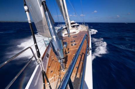 A sleek sailboat with a wooden deck cuts through the deep blue ocean, leaving a white wake under a clear sky.