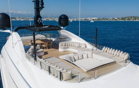 A luxury yacht deck with white lounge seating and a wooden dining area sits against a backdrop of deep blue ocean and a distant coastal city.