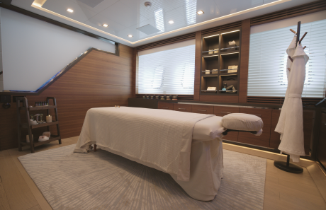 A white massage table sits on a light-colored rug in a modern spa room with wood-paneled walls, shelving, and blinds.