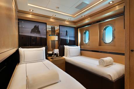 Two twin beds with white linens and rolled towels sit in a luxurious cabin featuring wooden walls, porthole windows, and modern ceiling lighting.