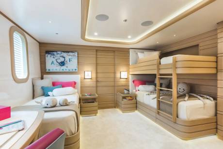 A modern bedroom features a single bed and a desk in the foreground, with bunk beds and light-wood paneled walls in the background.