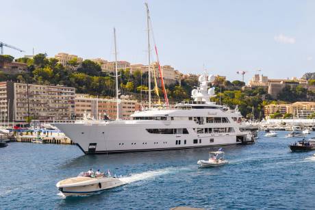 A massive white luxury yacht dominates the foreground of a blue harbor, with smaller boats cruising nearby and a hillside city with Mediterranean architecture in the background.