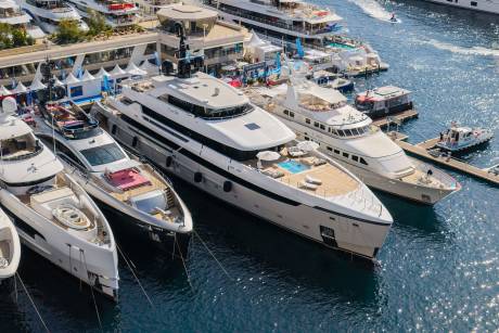 Several luxury yachts are docked side-by-side in a harbor, with a waterfront promenade and more boats visible in the background.
