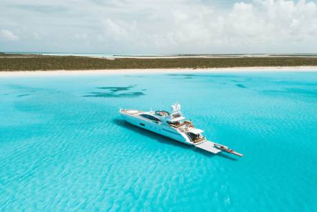 A luxury white yacht floats in crystal clear turquoise water with a sandy beach and green shrubbery in the background.