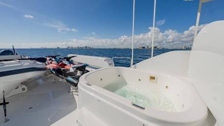 A white hot tub and jet skis sit on the deck of a boat overlooking a blue ocean and a distant city skyline under a sunny sky.