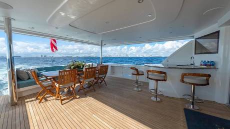 A luxury yacht deck features a dining area and bar in the foreground, overlooking a blue ocean and a distant city skyline in the background.