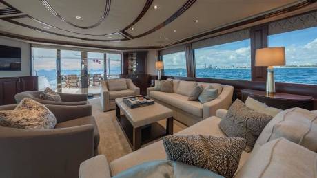 A luxurious yacht lounge with plush beige seating and a coffee table in the foreground opens up to a scenic ocean view and a distant city skyline in the background.