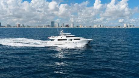 A white luxury yacht cruises through deep blue water with a city skyline and cloudy sky in the background.