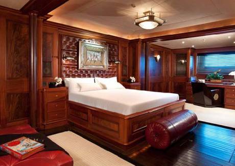 A luxurious bedroom with a large wooden bed and red bolster pillow in the foreground is set against a backdrop of rich wood-paneled walls and a built-in desk area.