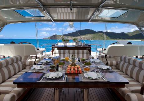 A set dining table with fruit and appetizers sits in the foreground of a luxury yacht, overlooking a turquoise ocean and lush green mountains in the background.