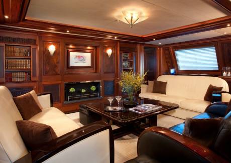 A luxurious wood-paneled lounge features white and brown leather sofas surrounding a dark coffee table in the foreground, with a fireplace and built-in bookshelves in the background.