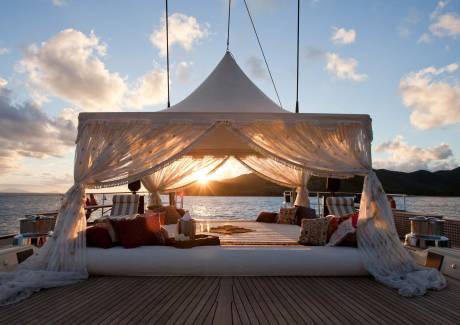 A luxurious white canopy bed with plush pillows sits on a wooden deck, set against a backdrop of calm ocean waters and distant mountains during a golden sunset.