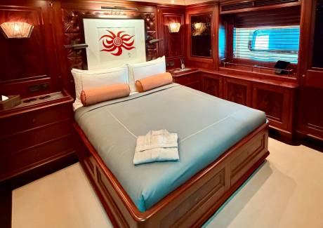 A bed with light blue linens and orange bolster pillows sits in a luxurious wood-paneled room featuring a red sun artwork and a window in the background.
