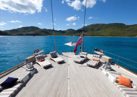 A wooden yacht deck with lounge chairs and a red flag sits in the foreground, set against a backdrop of turquoise water and lush green mountains under a blue sky.