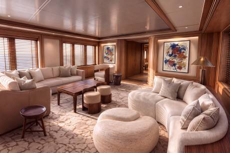 A luxurious lounge features plush white sofas and a large textured ottoman in the foreground, set against a background of warm wood paneling, abstract art, and slatted windows.