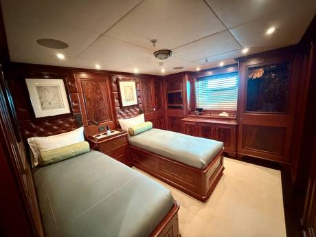 Two twin beds with light blue linens occupy the foreground of a luxurious wood-paneled cabin featuring a window and built-in cabinetry in the background.