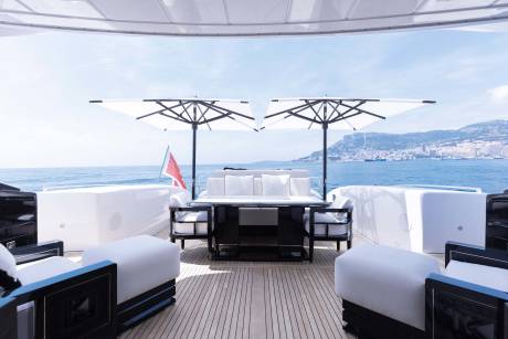 A luxurious yacht deck with white lounge furniture and umbrellas overlooks a blue sea and a distant coastal city.