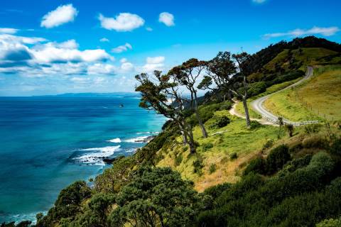 A winding road leads along a dramatic coastal cliffside overlooking the turquoise ocean under a bright blue sky.
