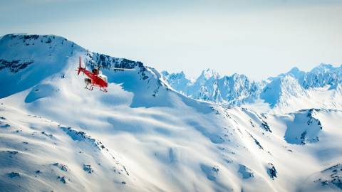 A bright red helicopter soars over a vast, snow-covered mountain range under a clear blue sky.