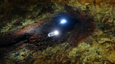 A suspended cage with bright lights descends into the colorful, rocky cavern of a dormant volcano.