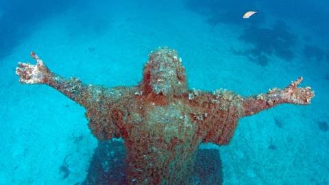 An underwater statue of Christ, encrusted with coral and seaweed, stands with arms outstretched in the clear blue ocean.