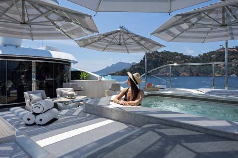 A woman in a black swimsuit and straw hat relaxes in a hot tub on a luxurious yacht with a stunning coastal backdrop.