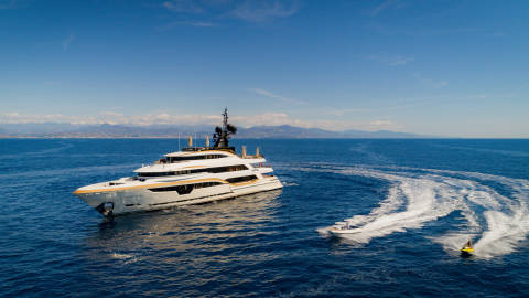 A sleek white and gold superyacht cruises on a calm blue sea, leaving a wake as a jet ski and a small boat speed nearby.