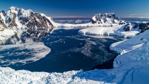 A majestic ship navigates the icy waters of Antarctica, surrounded by towering snow-capped mountains and floating icebergs under a clear blue sky.