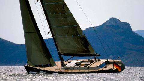 A sleek German yacht glides across the sparkling water with a mountainous coastline in the background.